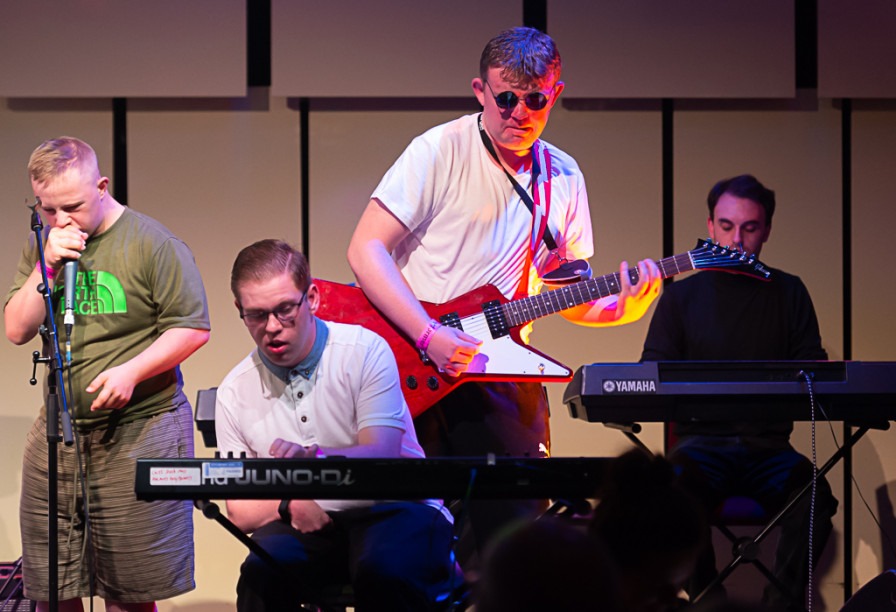 A young man is on stage playing a red guitar and wearing sunglasses. There is another man playing keyboard, another signing and another keyboard player in the background