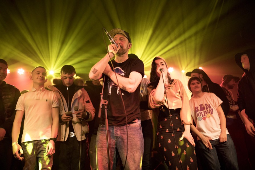 A crowd of young people stand on stage with microphones, backlit atmospherically. 
