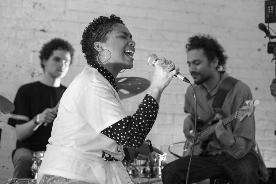 a young woman passionately sings into a microphone. her hair is short and curly and there are two band members seen in the background.