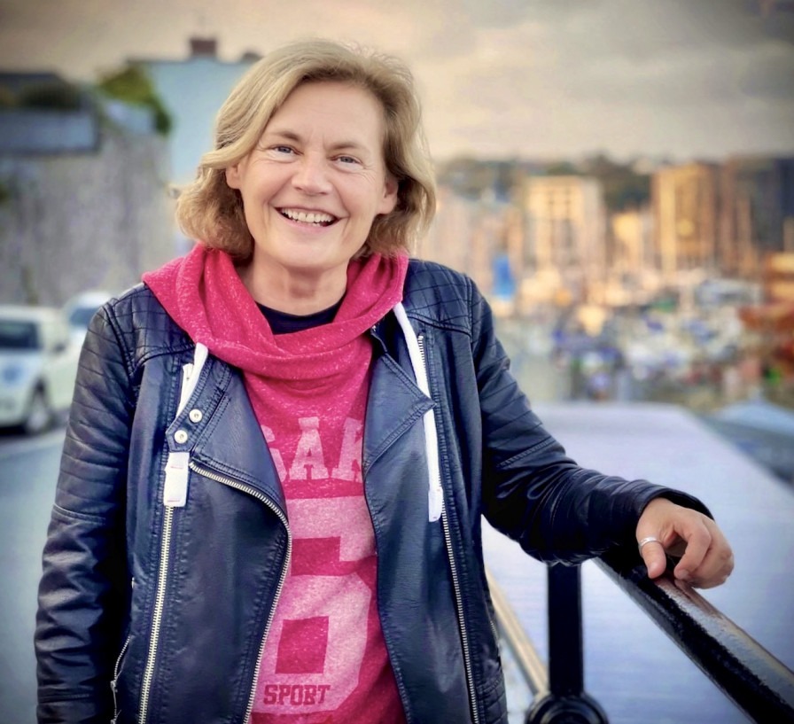 Woman with shoulder length brownish hair smiling wearing a bright red hooded top and leather jacket. She is leaning on a railing near a harbour in Plymouth at sunset 