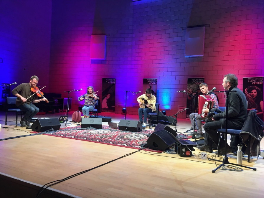 Five musicians seated on stage playing fiddle, viola, guitar, piano accordion and concertina. 