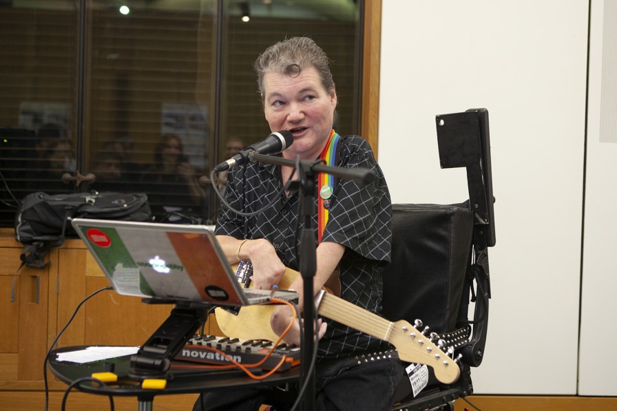 John Kelly performs, sitting in front of his microphone and laptop with his adapted Kellycaster guitar round his neck on a rainbow strap