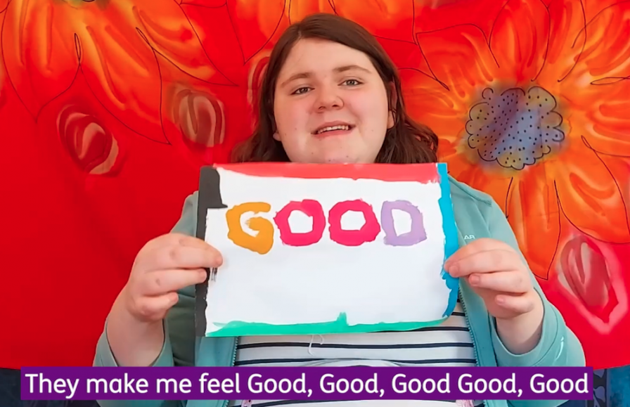 A girl with brown hair sits against a backdrop with large red flowers holding a hand-painted colourful sign reading 'good' in capital letters. Underneath there are captions reading 'they make me feel good, good, good, good, good.'