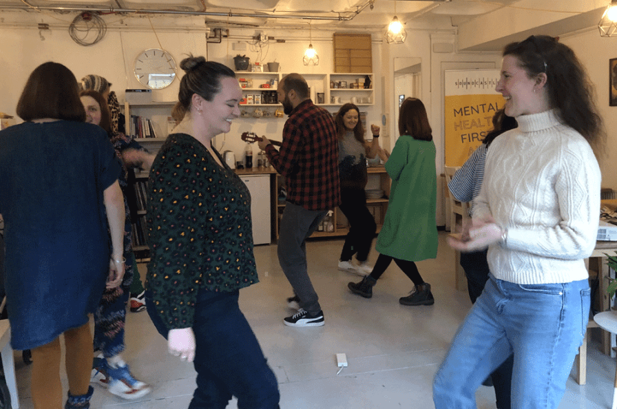 A room of people smiling and dancing - someone in the middle has a ukulele, and there's a poster saying Mental Health First Aid in the background